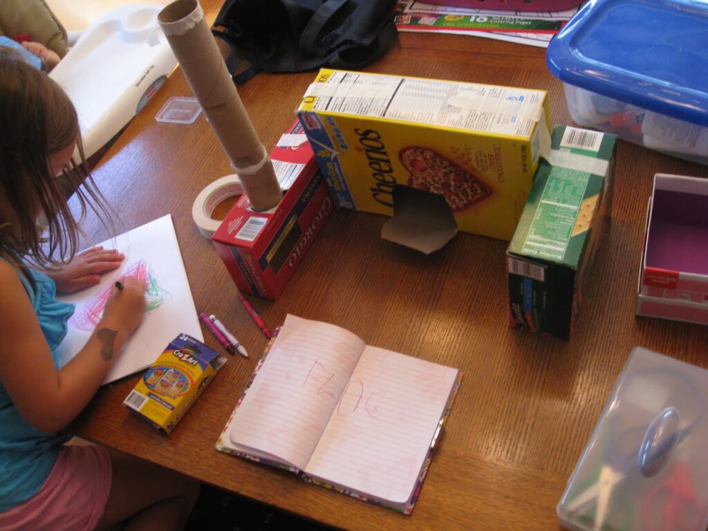 Child writing on paper by cardboard creation made from old cereal box, cardboard tube, and cracker boxes