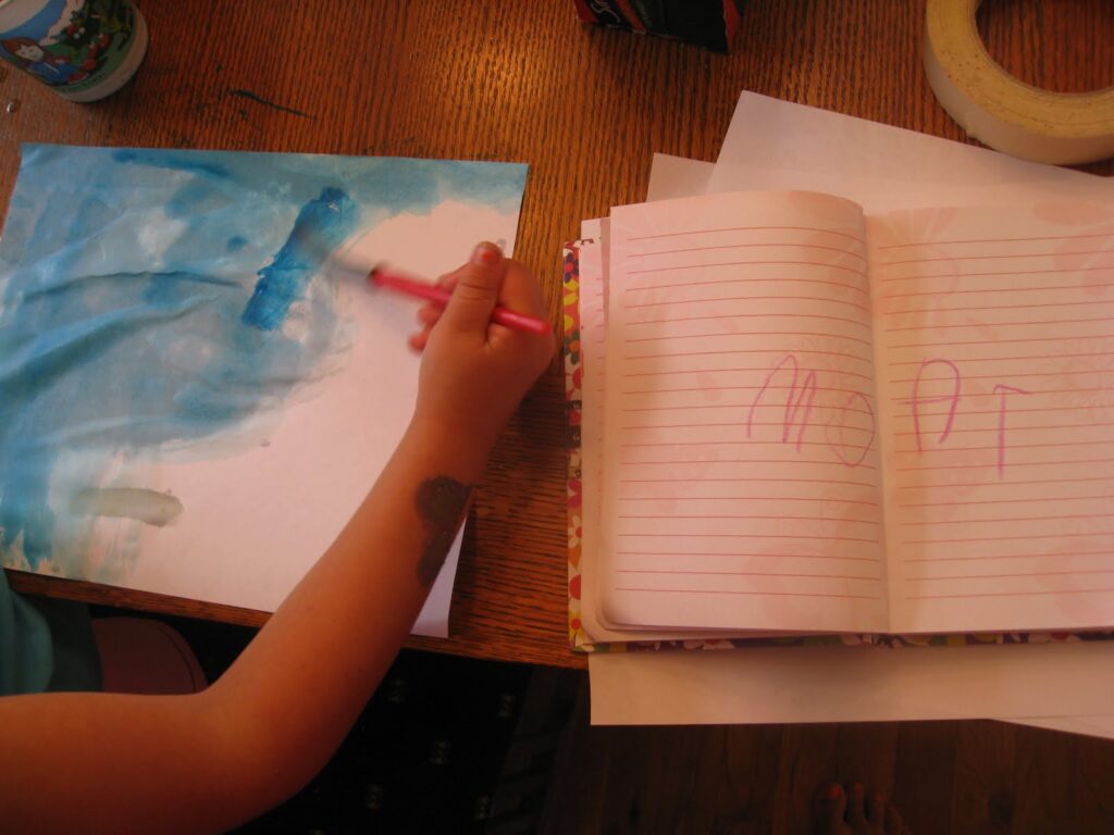 Child painting a paper blue and a notebook with "moat" written in child's writing 