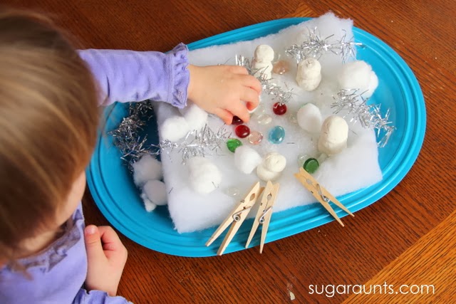 Child reaching for items in a Frosty the snowman sensory bin with silver tinsel, baking soda dough snowmen, cotton balls, clothes pins, and red and green gems.