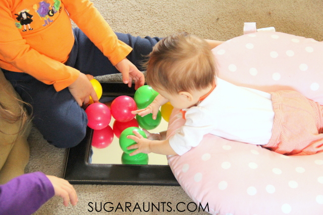 baby laying on her belly on a nursing pillow, playing with ball pit balls on a mirror while older siblings help