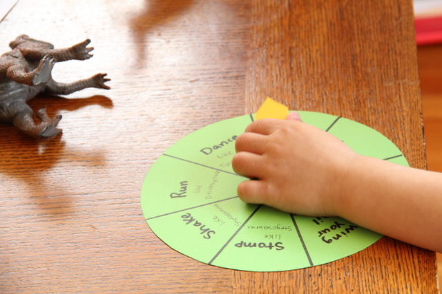 Child's hand spinning a game spinner  for a dinosaur game with words for gross motor skills: stomp, shake, run, dance.