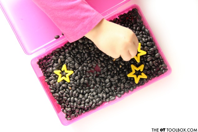 sensory bin in a pencil case with black beans as a sensory base and yellow stars. Child's hand reaches for a yellow star