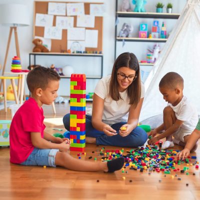 Hermosa maestra y niños pequeños jugando con bloques de construcción alrededor de un montón de juguetes en el jardín de infantes